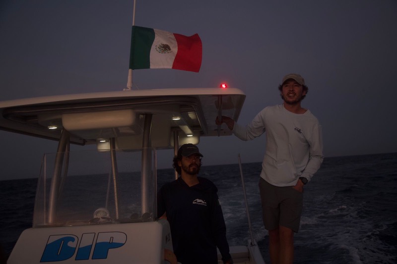 Matías, in dark clothes, at the helm of La Dip and Eduardo, in light clothes, up top at dusk under the Mexican flag