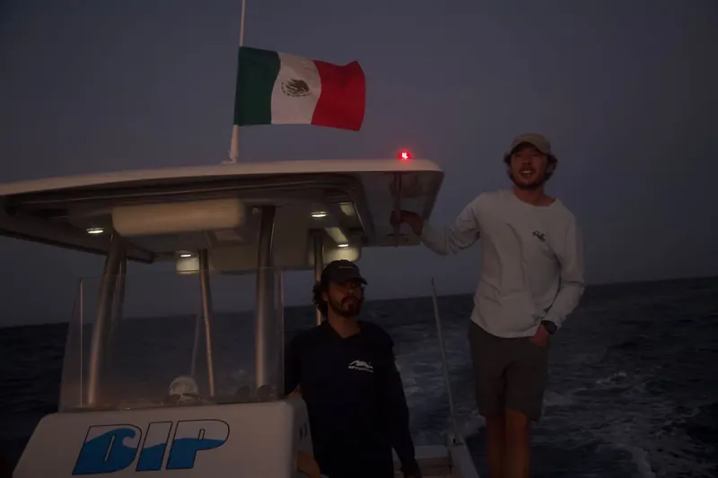 Matías, in dark clothes, at the helm of La Dip and Eduardo, in light clothes, up top at dusk under the Mexican flag